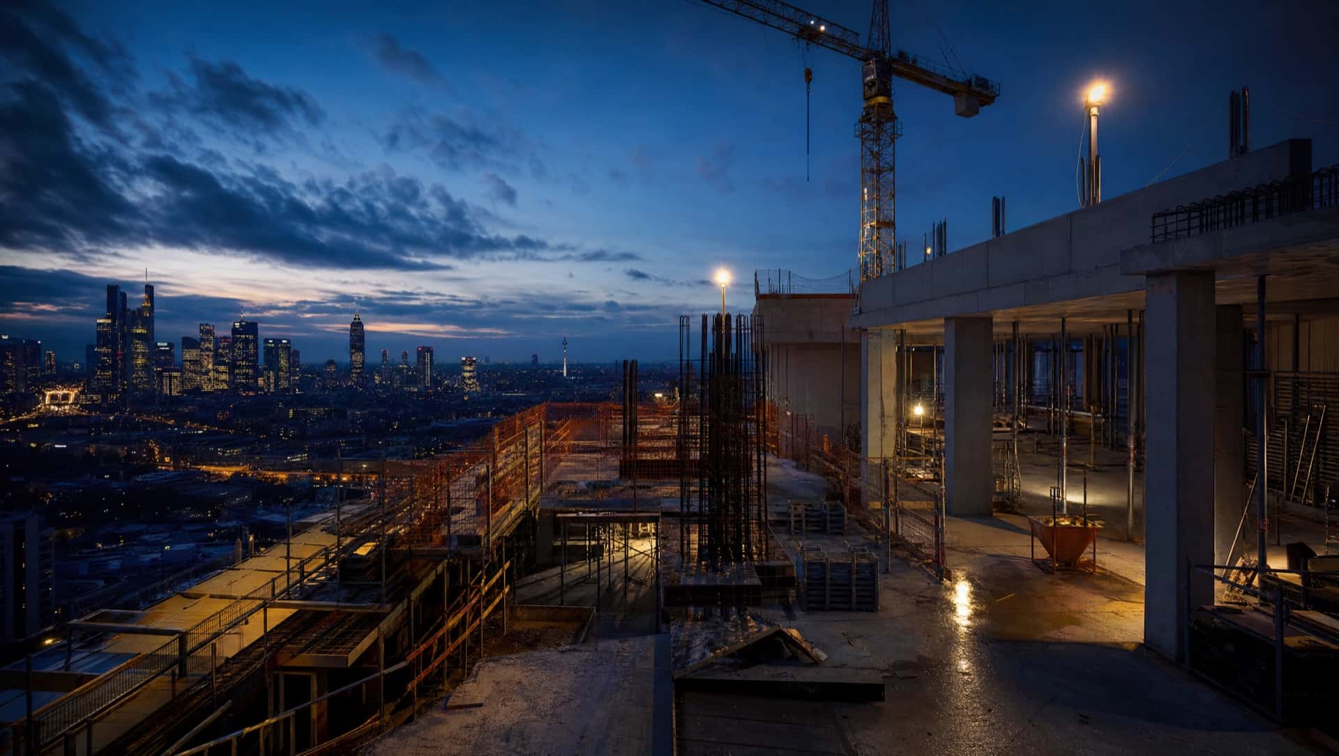 Aktive Hochbau-Baustelle mit Bewehrung, Schalung und Kran vor der Frankfurter Skyline zur blauen Stunde