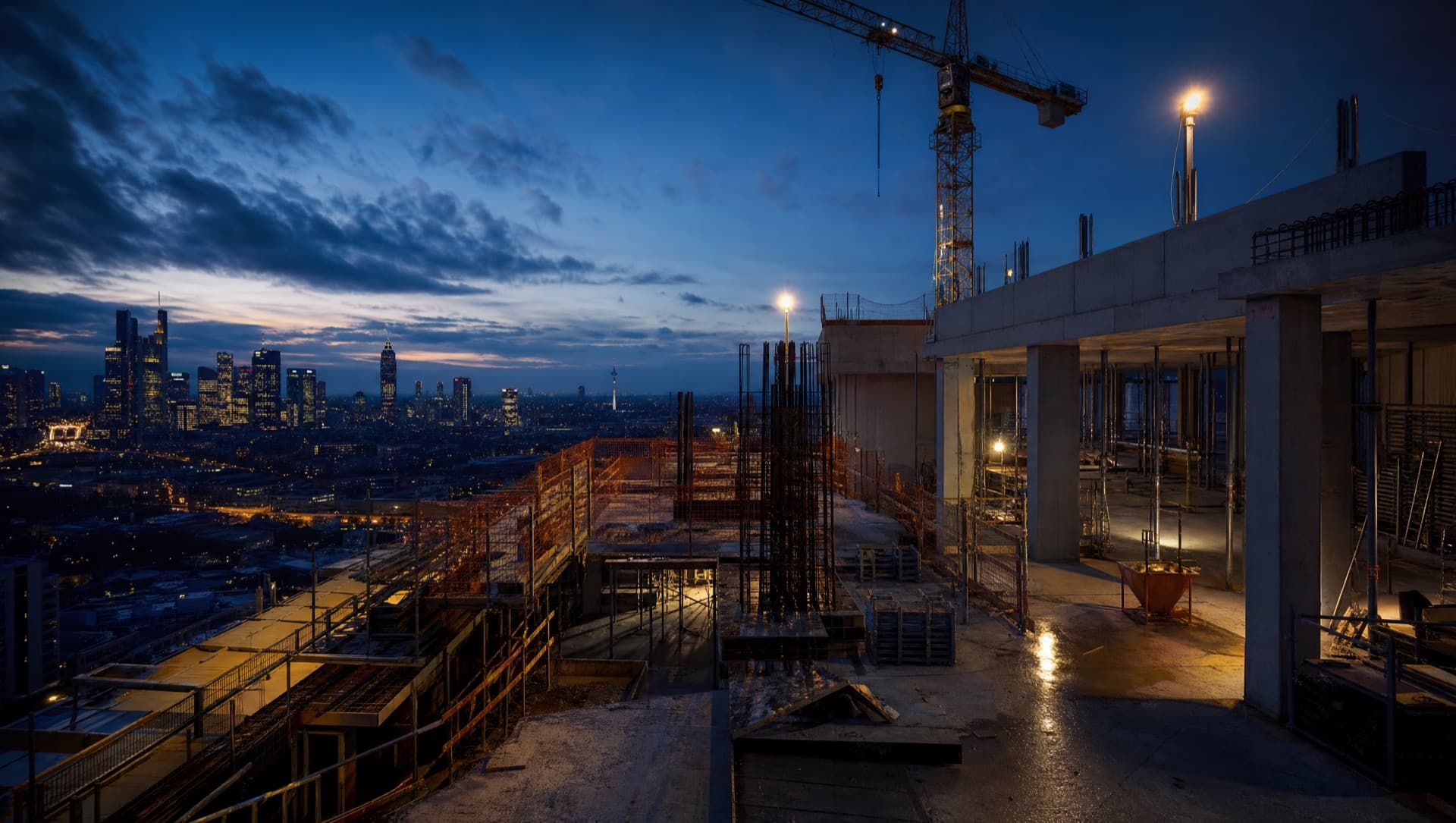 Aktive Hochbau-Baustelle mit Bewehrung, Schalung und Kran vor der Frankfurter Skyline zur blauen Stunde