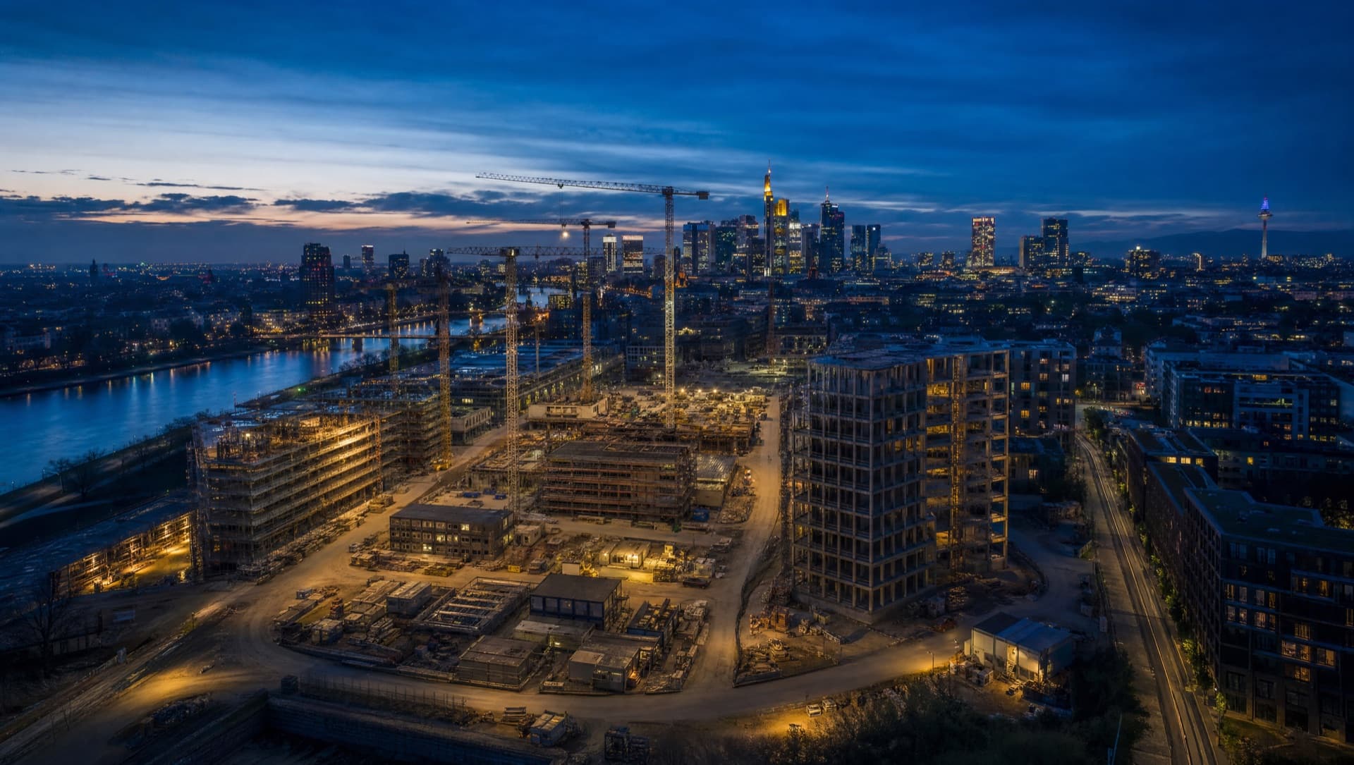 Luftaufnahme mehrerer Bauvorhaben mit Kränen vor der Frankfurter Skyline zur blauen Stunde
